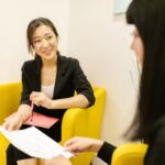 Two professional women having a discussion in an office with documents in hand.